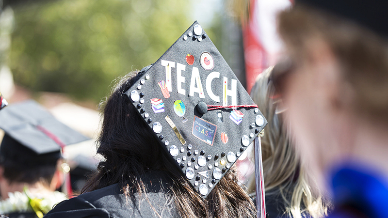 A black graduation cap decorated with teaching stickers and white letters that say teach